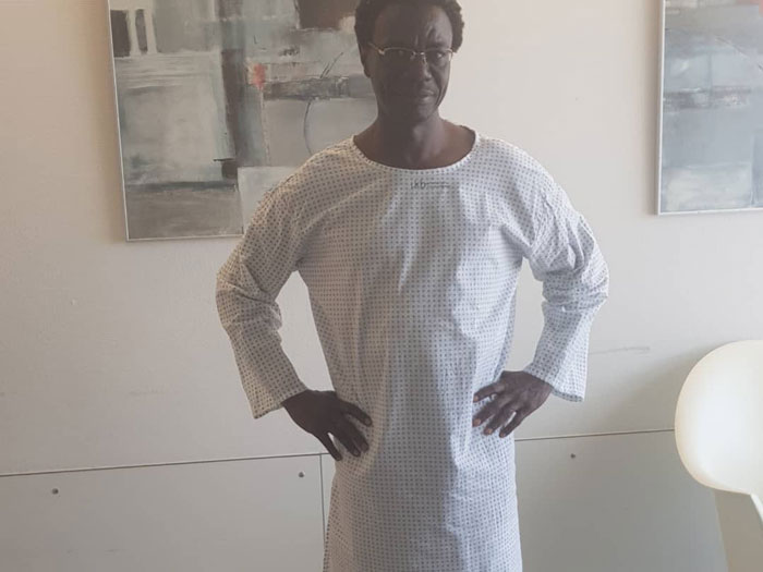 Ein farbiger Mann posiert mit weißem Nachthemd im Krankenzimmer